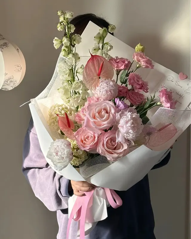 A soft pink bouquet of roses, peonies, lisianthus and anthurium in white wrap with a handwritten card